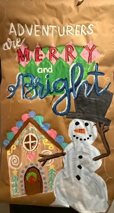 Hand-painted holiday poster on kraft paper with 'Adventurers are Merry and Bright' lettering in white, red, and blue, a snowman in a top hat, and a candy-covered gingerbread house