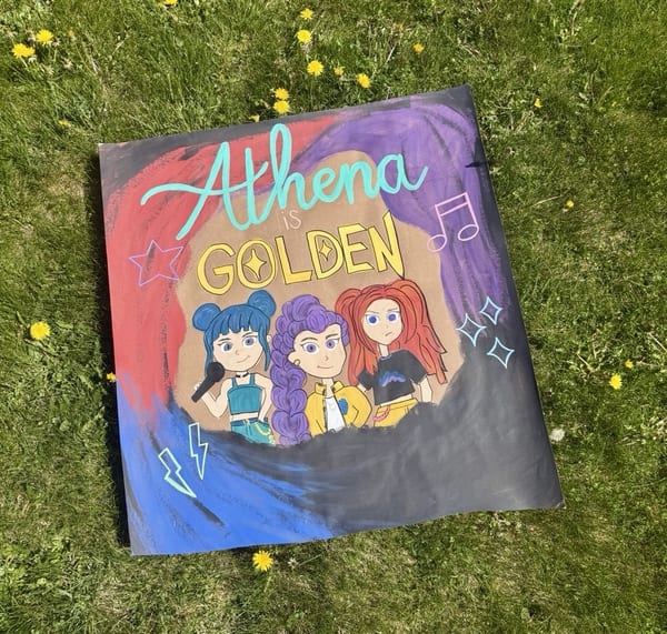 Hand-painted character banner on kraft paper with turquoise 'Athena' script, yellow 'is Golden' lettering, three colorful pop singers, music notes, stars, sparkles, and sweeping red, purple, and blue paint