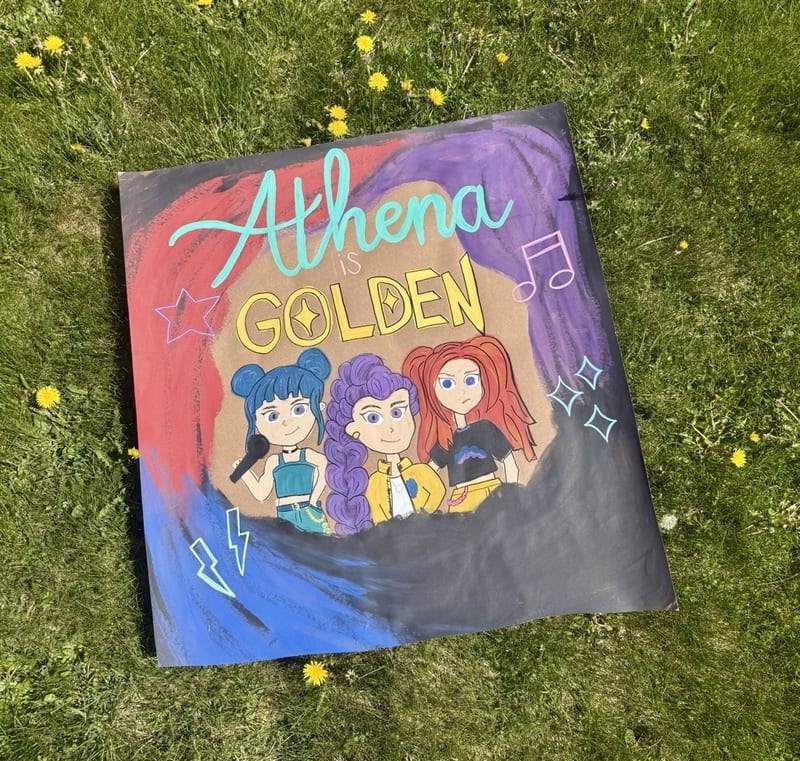 Hand-painted character banner on kraft paper with turquoise 'Athena' script, yellow 'is Golden' lettering, three colorful pop singers, music notes, stars, sparkles, and sweeping red, purple, and blue paint