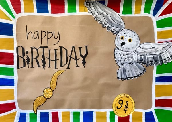Birthday banner on brown kraft paper with black 'Happy Birthday' lettering, a flying gray owl, a winged golden ball, a round train-platform ticket, and a colorful striped border