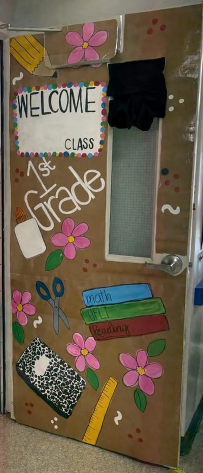Classroom door with WELCOME, CLASS, and 1st Grade lettering on a brown background, pink flowers, colorful dots, and illustrated school supplies