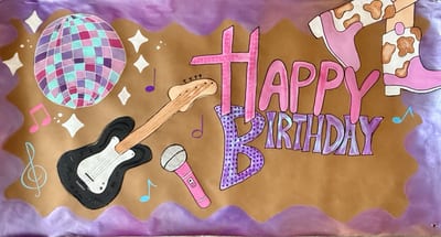 Birthday banner on kraft paper with pink and purple 'Happy Birthday' lettering, a disco ball, black electric guitar, pink microphone, cow-print boots, music notes, and a purple border