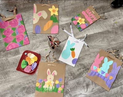 Six hand-painted Easter gift bags on brown and white paper with colorful bunnies, carrots, eggs, spring flowers, pink lettering, and ribbon handles, arranged around paint supplies on a wood-look floor
