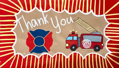 Hand-painted thank you banner for firefighters on kraft paper with white script lettering, a red and blue firefighter Maltese cross logo, a cartoon red fire truck with extended ladder, and a bold red and yellow sunburst border with white scalloped frame