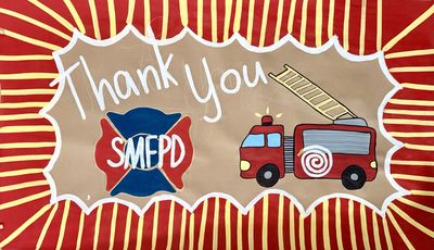 Hand-painted thank you banner for SMFPD on kraft paper with white script lettering, a red and blue firefighter maltese cross logo, a cartoon red fire truck with extended ladder, and a bold red and yellow sunburst border with white scalloped frame