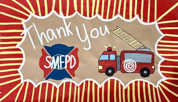 Hand-painted thank you banner for SMFPD on kraft paper with white script lettering, a red and blue firefighter maltese cross logo, a cartoon red fire truck with extended ladder, and a bold red and yellow sunburst border with white scalloped frame