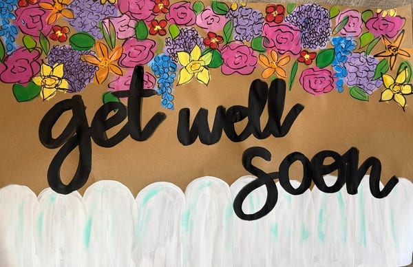 Hand-painted get well soon banner on kraft paper with bold black script lettering, pink roses, purple lilacs, yellow daffodils, and orange blooms across the top, and a white picket fence along the bottom