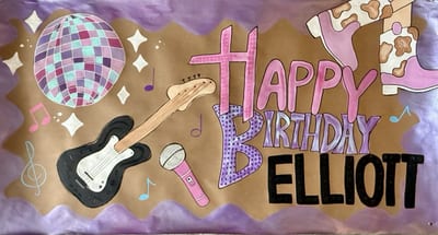 Hand-painted birthday banner on brown kraft paper with pink, purple, and black 'Happy Birthday Elliott' lettering, a disco ball, black electric guitar, pink microphone, cow-print cowgirl boots, colorful music notes, and a wavy purple border