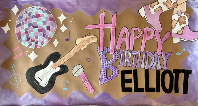 Hand-painted birthday banner on brown kraft paper with pink, purple, and black 'Happy Birthday Elliott' lettering, a disco ball, black electric guitar, pink microphone, cow-print cowgirl boots, colorful music notes, and a wavy purple border