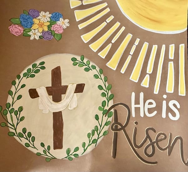 Hand-painted Easter banner on brown kraft paper with a draped wooden cross inside a cream circle, green leafy branches, colorful flowers, yellow sun rays, and white and brown 'He is Risen' lettering