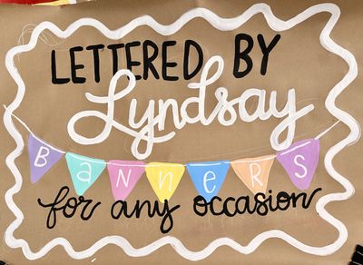 Hand-painted business banner on brown kraft paper with 'Lettered by Lyndsay' in black block letters and white script, colorful triangle pennant flags spelling 'Banners,' and 'for any occasion' in black script, all framed by a white wavy border