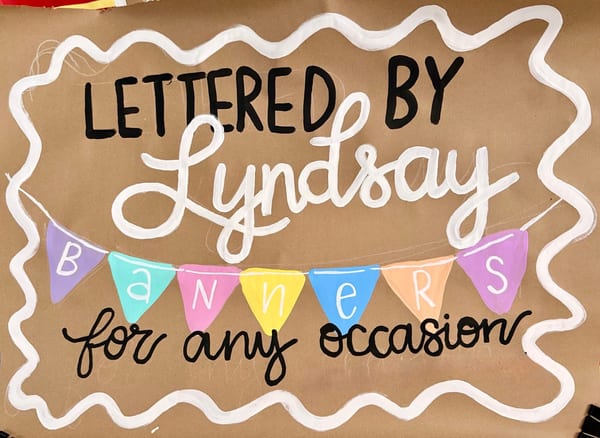 Hand-painted business banner on brown kraft paper with 'Lettered by Lyndsay' in black block letters and white script, colorful triangle pennant flags spelling 'Banners,' and 'for any occasion' in black script, all framed by a white wavy border