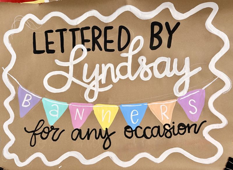 Hand-painted business banner on brown kraft paper with 'Lettered by Lyndsay' in black block letters and white script, colorful triangle pennant flags spelling 'Banners,' and 'for any occasion' in black script, all framed by a white wavy border