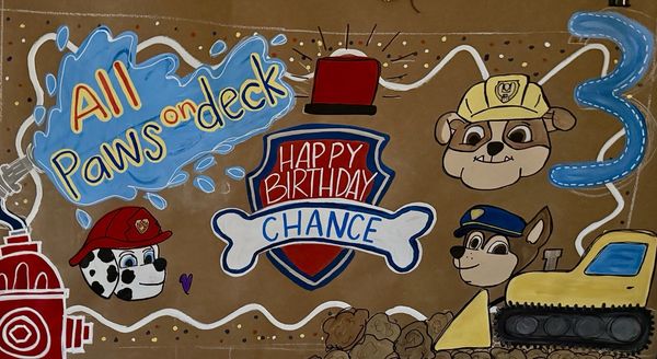 Hand-painted Paw Patrol birthday banner on kraft paper with 'All Paws on Deck' in blue and white lettering, a shield reading 'Happy Birthday Chance,' the number 3, and Marshall, Chase, and Rubble painted alongside their vehicles