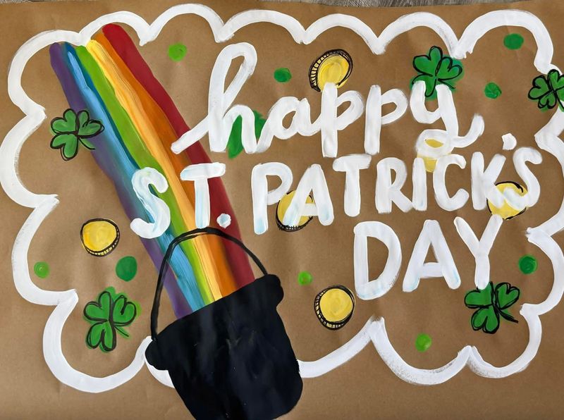 Hand-painted St. Patrick's Day banner on brown kraft paper with white script 'Happy St. Patrick's Day' lettering, a black pot of gold, a rainbow, scattered gold coins, green shamrocks, green polka dots, and a white scalloped border