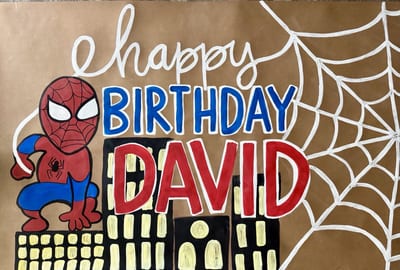 Hand-painted superhero birthday banner on brown kraft paper with a red-and-blue masked hero, white webbing forming the word 'happy,' bold blue and red lettering for 'Birthday David,' and a black city skyline