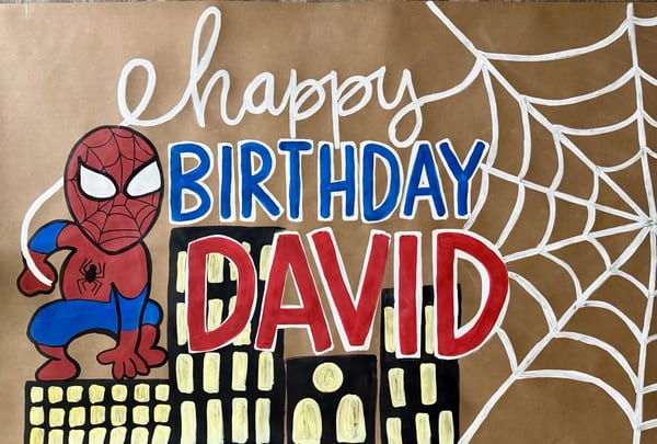 Hand-painted superhero birthday banner on brown kraft paper with a red-and-blue masked hero, white webbing forming the word 'happy,' bold blue and red lettering for 'Birthday David,' and a black city skyline