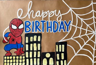 Superhero birthday banner on kraft paper with a red-and-blue masked hero, white webbing that spells 'happy,' bold blue 'BIRTHDAY' lettering, and a black city skyline